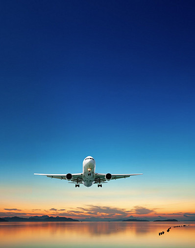 An airplane flying low over calm water during a colorful sunset with mountains in the distance