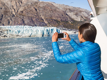 A person standing on a cruise ship deck photographing a glacier and icy blue water with rocky mountains in the background.