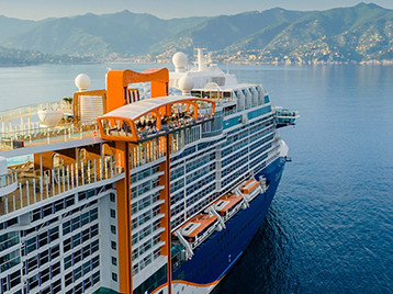 A modern cruise ship sailing along a calm coastline with mountains in the background under a clear blue sky.