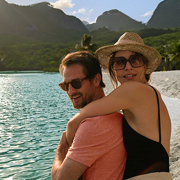 Two people sitting on a tropical beach beside turquoise water with lush green mountains in the background