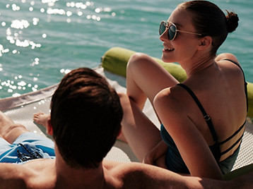 Two people relaxing on a lounge at the edge of the water on a sunny day.