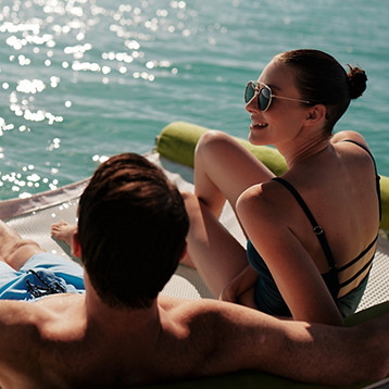 Two people relaxing on a lounge at the edge of the water on a sunny day.