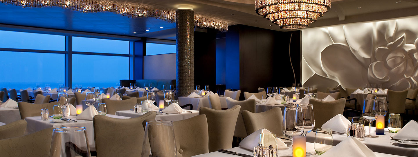 An upscale restaurant with a twinkling chandelier, white table linens and windows overlooking the ocean.