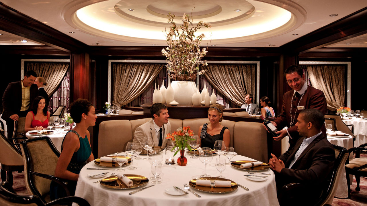 A group of diners seated at a large table in a fine dining restaurant are shown a bottle of wine by a waiter in uniform.