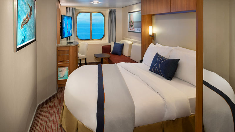 Celebrity Cruises Deluxe Ocean View Stateroom, featuring a cream leather couch, Queen size bed, small coffee table and desk.