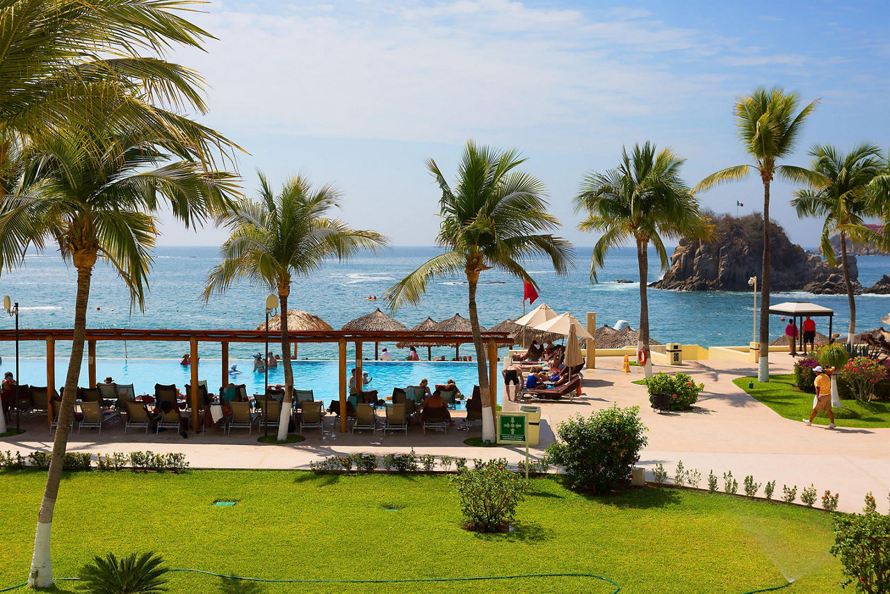 Palm trees along the beach with tiki huts and beach-goers enjoying the day in Huatulco, Mexico