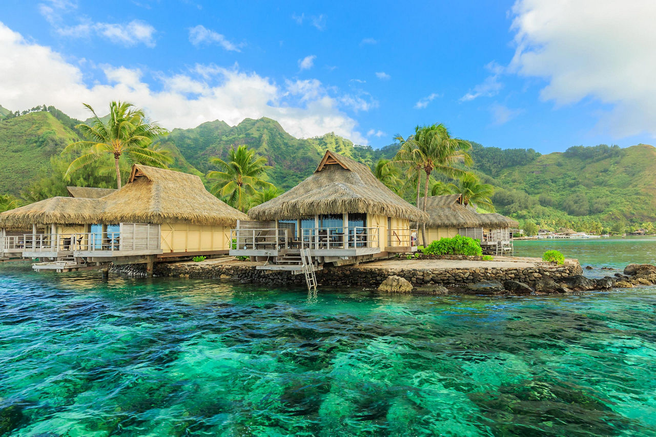 Tiki huts sitting on top of beautiful crystal turquoise waters with mountains in the background in Papeete, Tahiti
