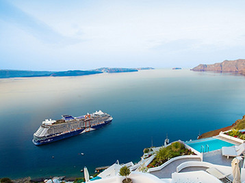 A cruise ship sailing across calm blue water with white coastal buildings and dramatic island cliffs overlooking the sea.