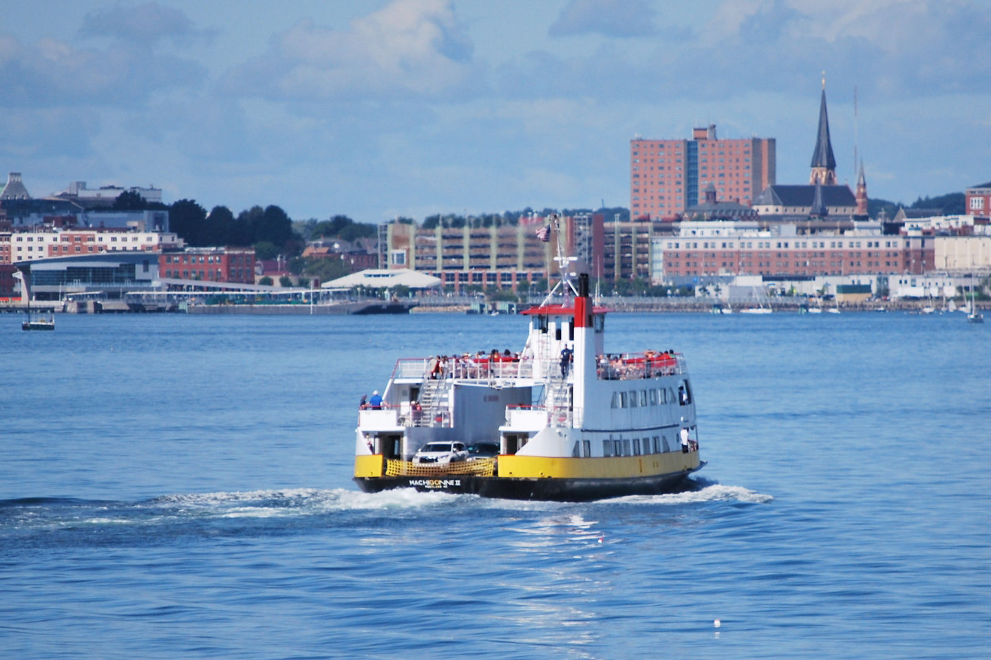 A Casco Bay Lines ferry moves across Portland’s harbor, with calm water and city buildings behind it. - Portland, Maine