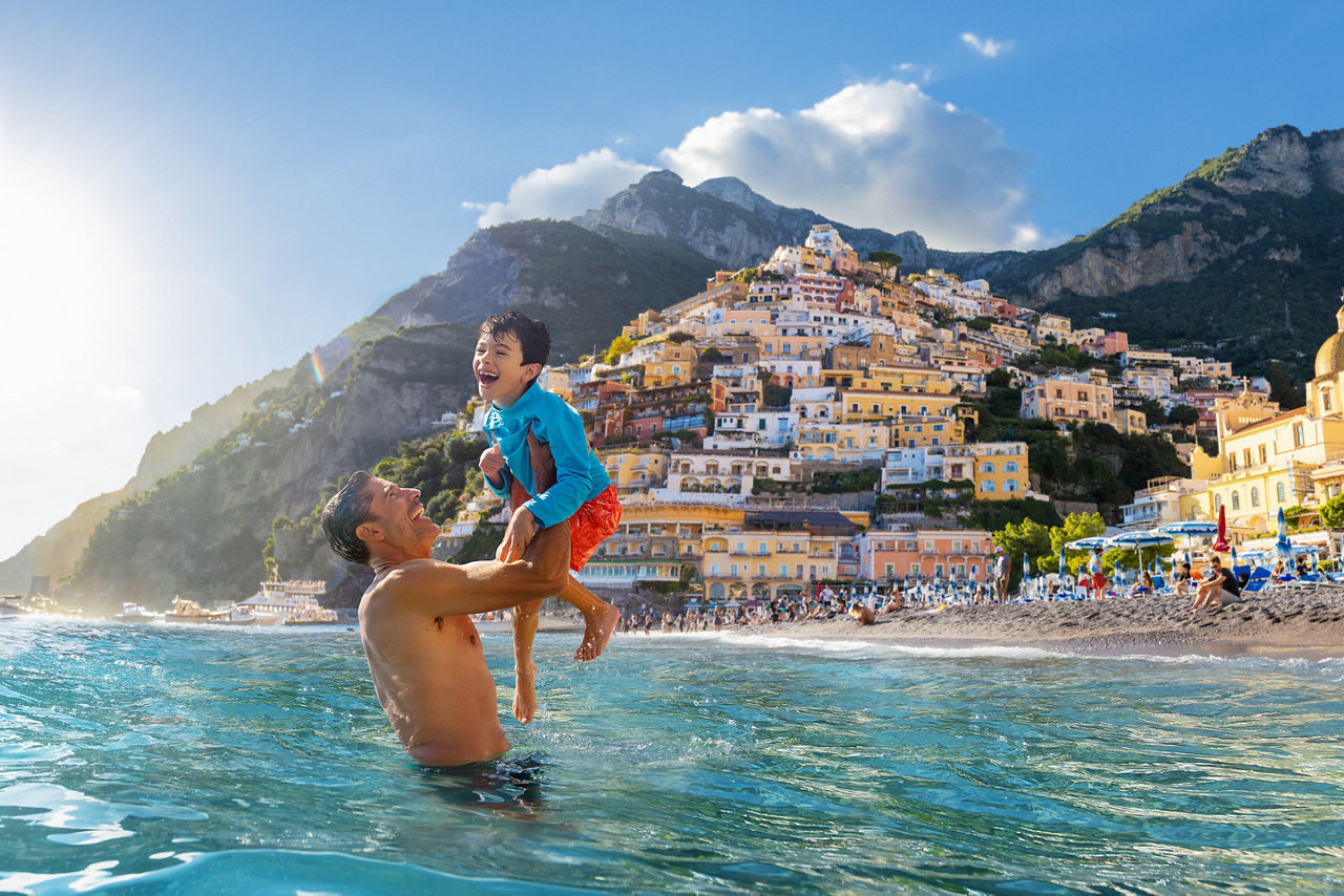 A father lifts his son while standing in clear blue water at a busy beach in Positano, Italy