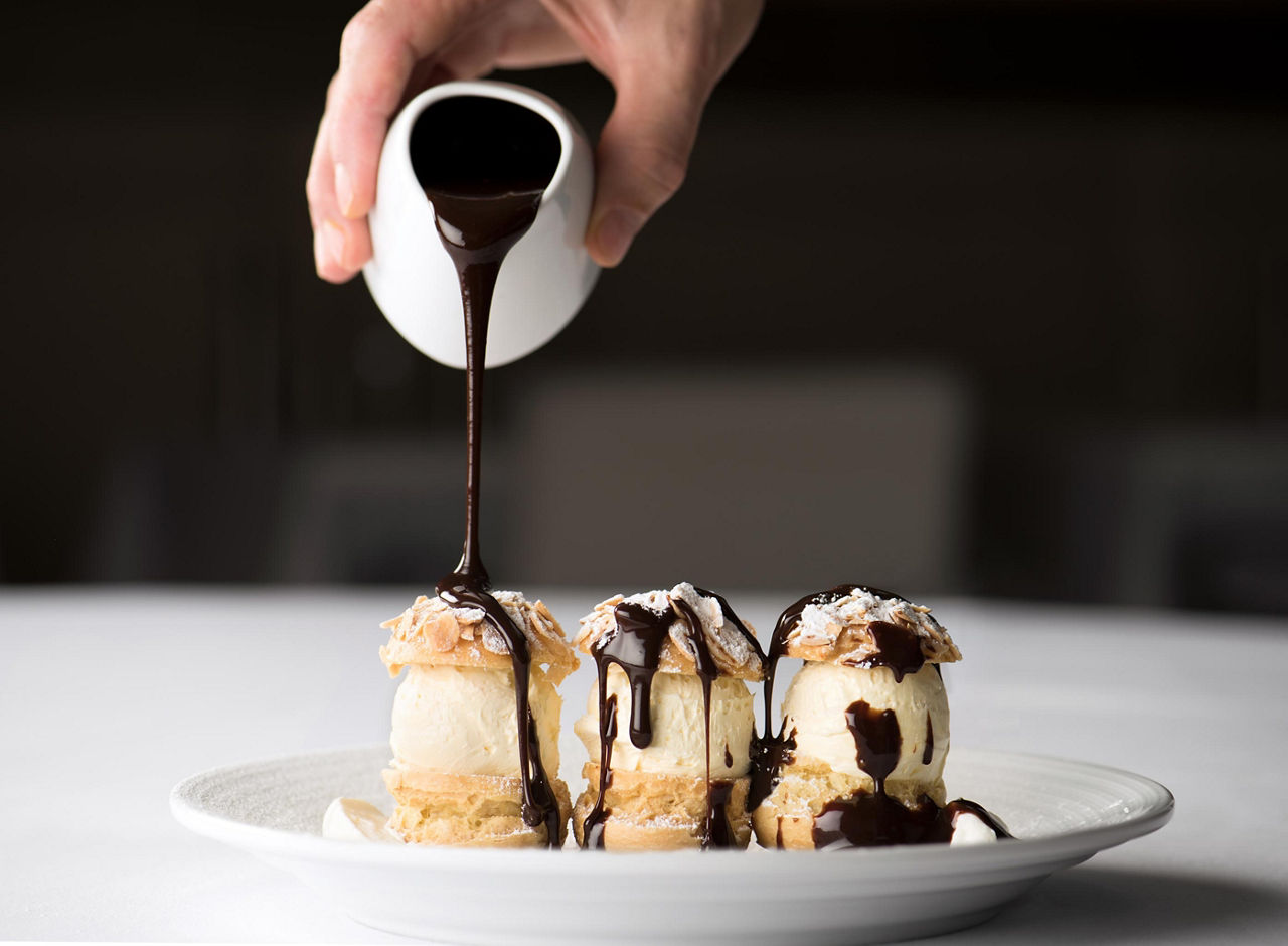 A hand pouring chocolate sauce over a vanilla ice cream pastry at the Main Dining Room