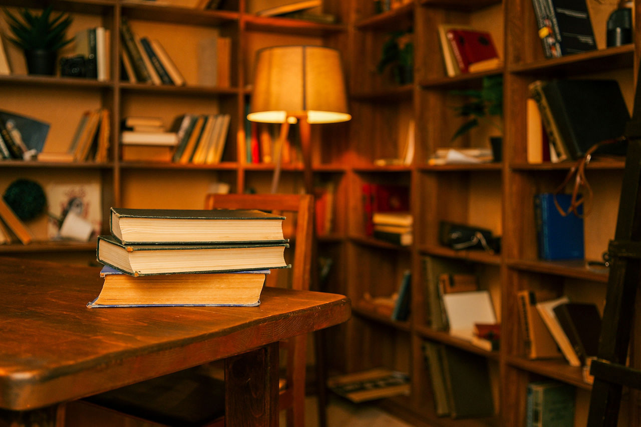 Cozy library interior with stacked books on a wooden table and warm lamp light