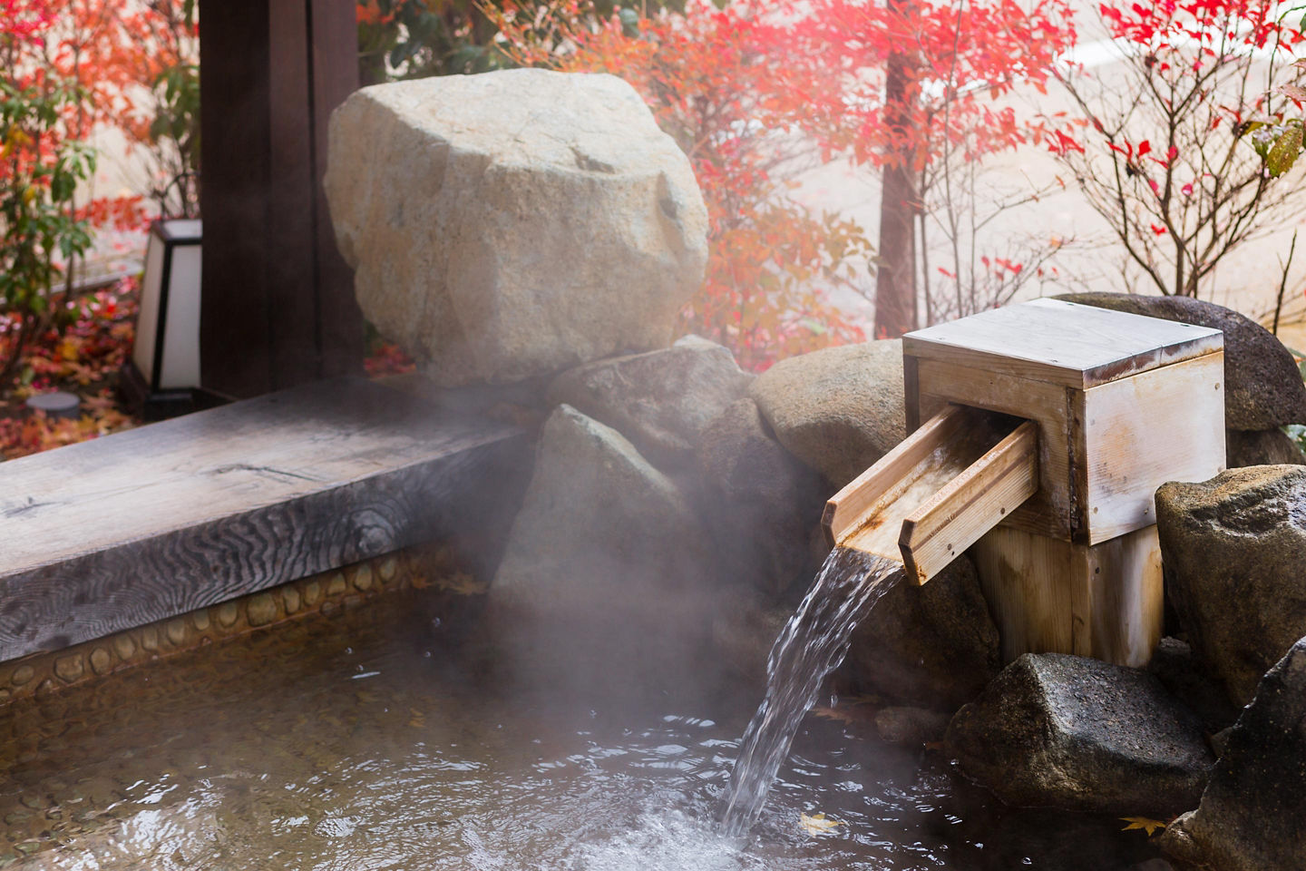 Steam rises from a serene Japanese hot spring bath surrounded by rocks and warm natural light. - Kobe, Japan