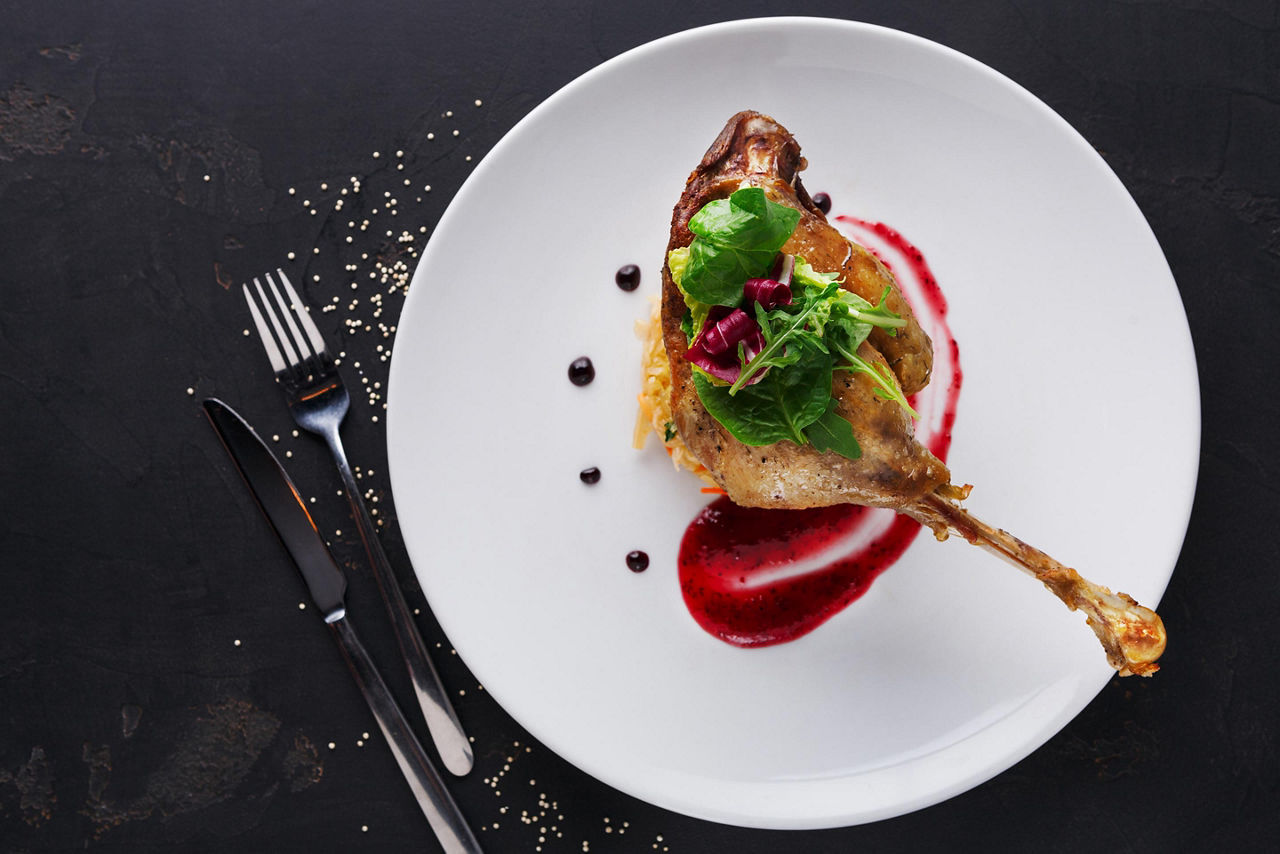 Duck confit with braised cabbage, baked apple and cranberry sauce served on snow white plate with cutlery on black table background