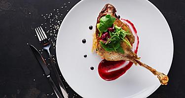 Duck confit with braised cabbage, baked apple and cranberry sauce served on snow white plate with cutlery on black table background