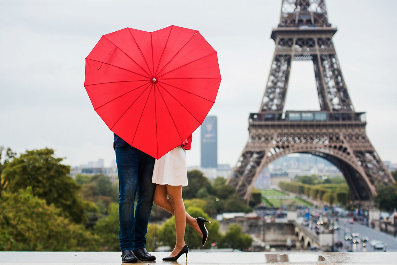 Couple visiting the Eiffel Tower standing with a red heart-shaped umbrella in Paris. Europe.