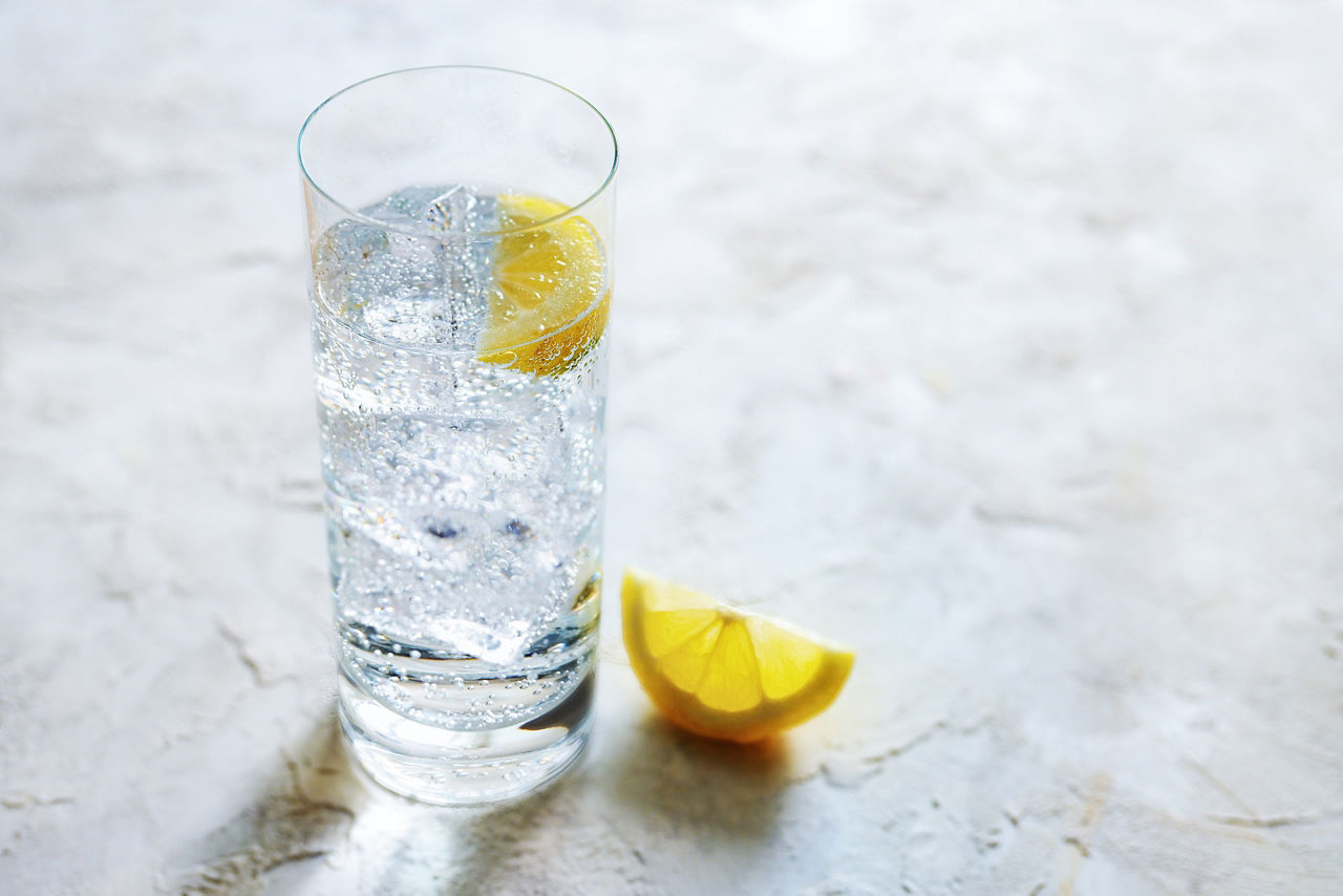 Fresh carbonated mineral water with ice cubes and lemon slices in a drinking glass, refreshing drink on a light stone background, copy space, selected focus