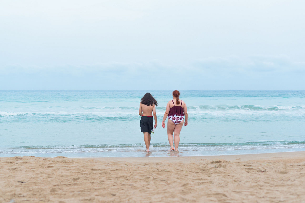 Boy with long hair and bermuda shorts and a young adult entering the sea for a swim. 