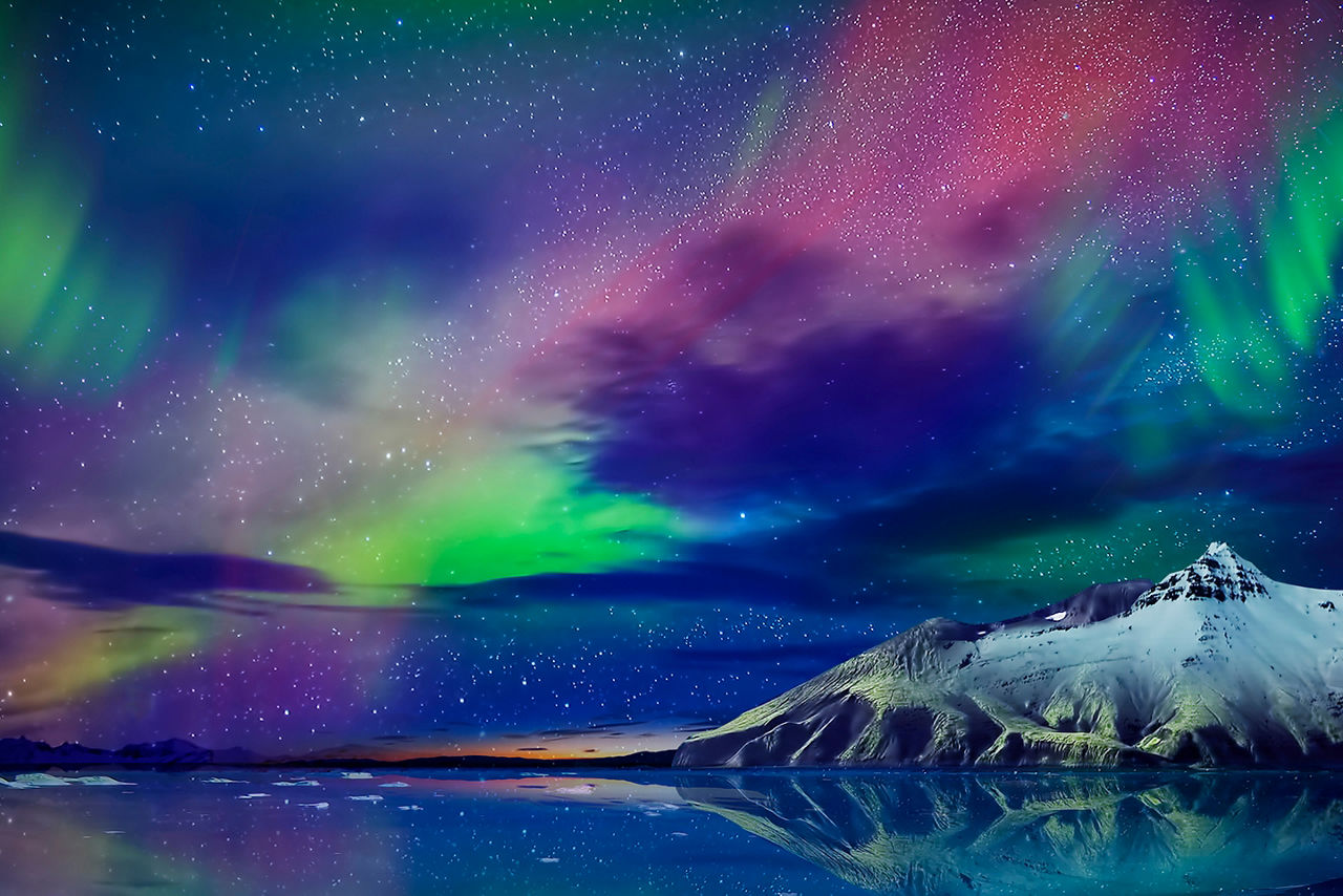 Gorgeous, unreal beautiful night view of the reflection of the northern lights in the water of the ocean and snow-capped mountains.