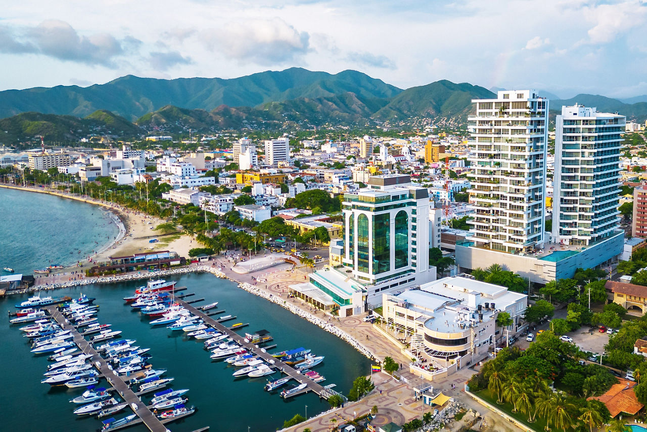 Drone view of Santa Marta Marina and El Rodadero district in Santa Marta, Colombia, with sailboats, waterfront developments, Caribbean coastline, and the Sierra Nevada de Santa Marta mountains creating a dramatic backdrop.