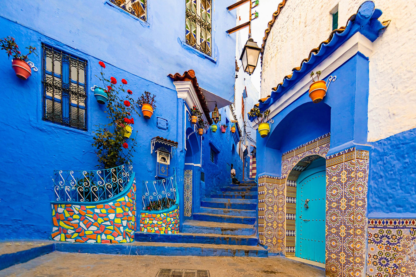 A narrow Chefchaouen street glows with vivid blue walls, colorful pots, and bright sunlight. - Chefchaouen, Morocco