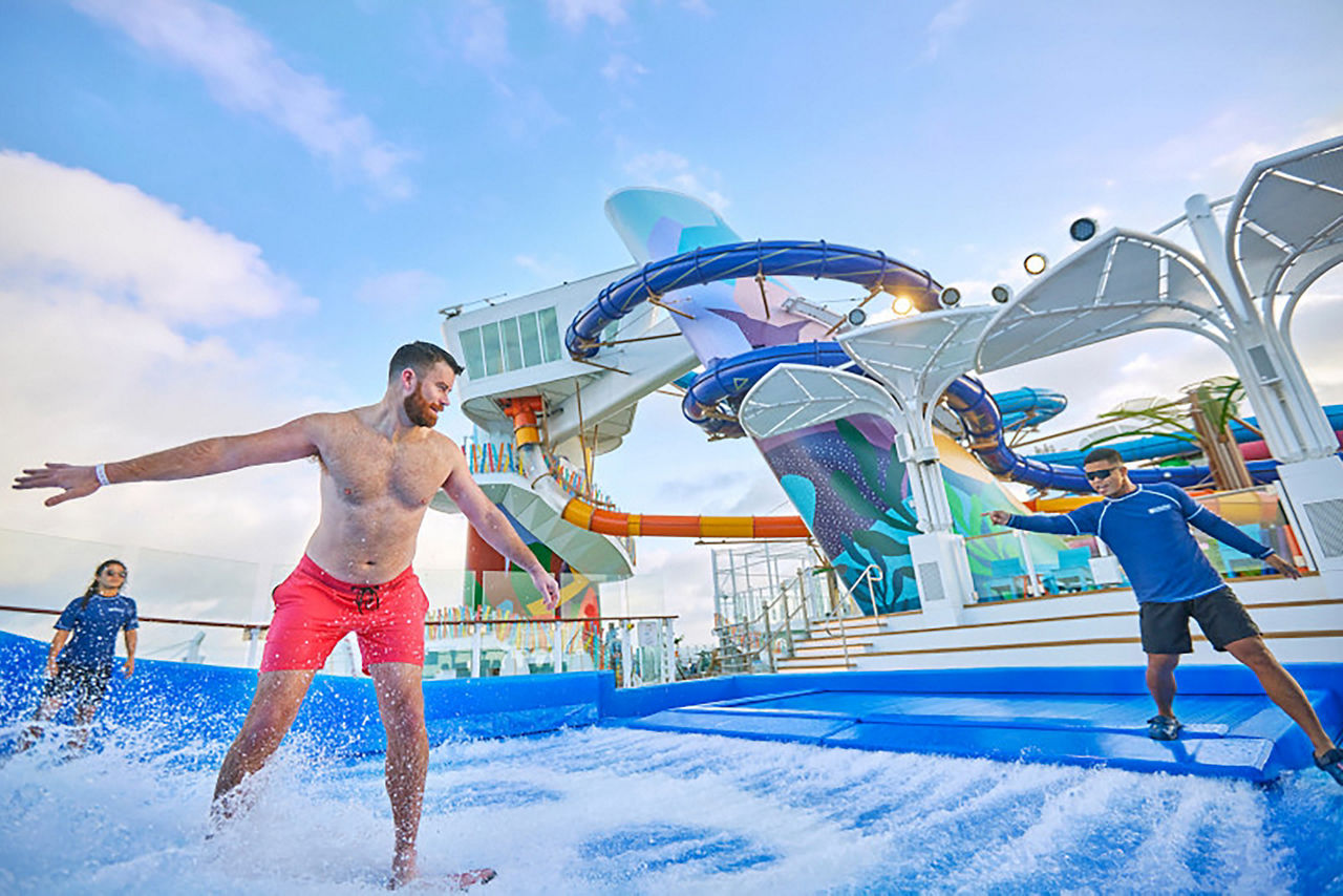 Man riding surfboard and staff attendants at side giving instructions on the flowrider