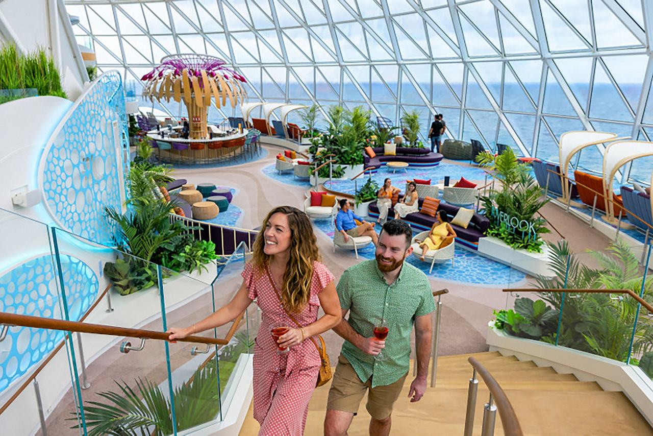 Couple walking up stairs with view of Overlook behind them on Icon of the Seas, ocean horizon seen through windows,