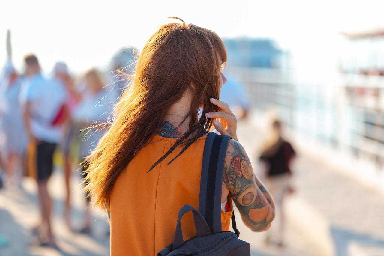 Rear view of young woman in orange dress with tattoos wearing a backpack walking down the port. 
