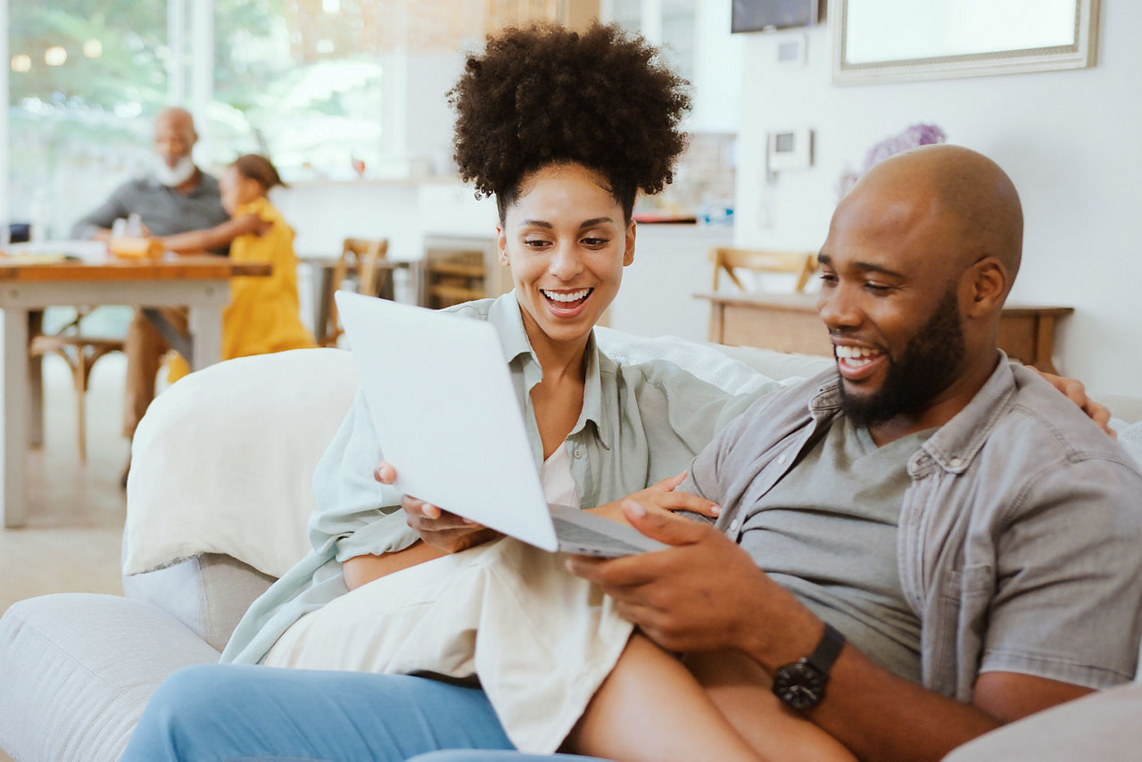 Couple At Home Using Laptop To Shop Or Book Holiday With Multi-Generation Family In Background