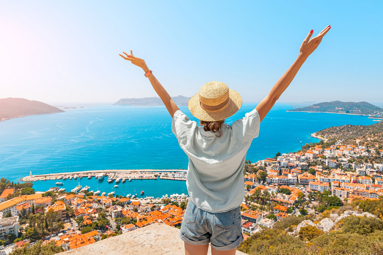 Happy woman with open arms stands on the viewpoint and enjoys the panorama of Kas resort town of the Mediterranean sea in Turkey